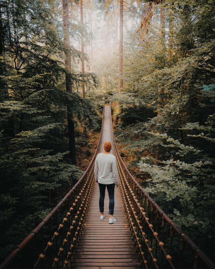 A woman walking alone on a bridge surrounded by tall trees, with sunlight shining through the forest ahead.